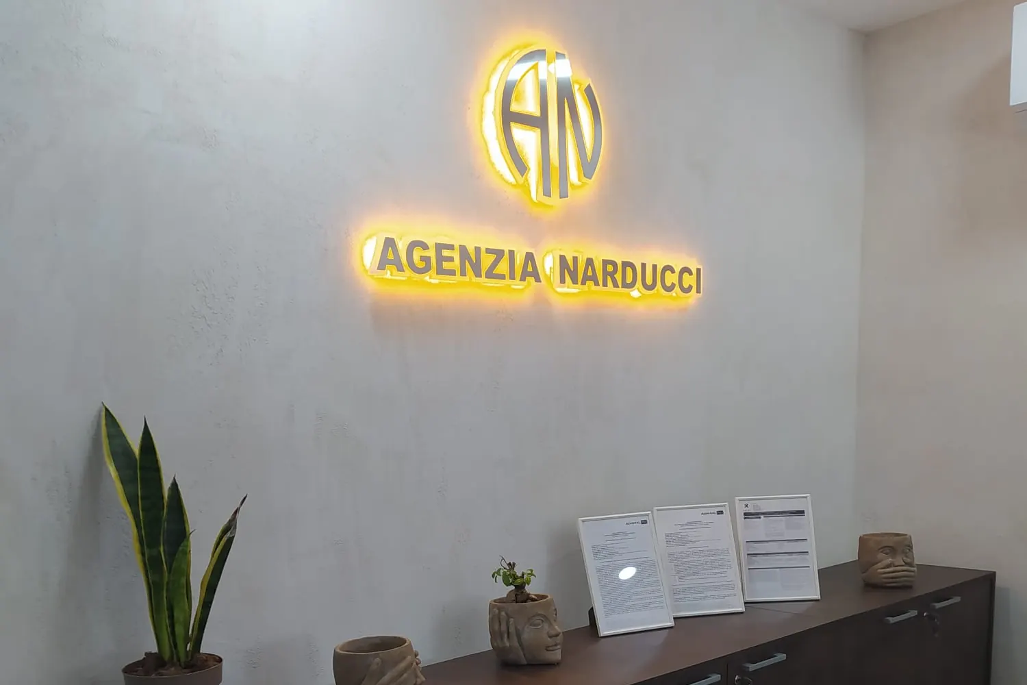 La parete dell'Agenzia Narducci col logo illuminato, sopra una credenza con delle piante
